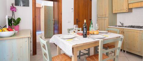 A dining area with a table set for two, chairs with woven seats, a bowl of fruit, and a bottle of wine on the table.
