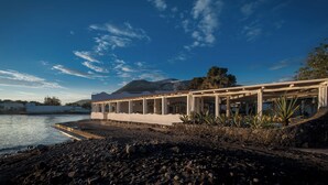 A coastal building with a stone foundation, a flat roof, and a series of columns supporting a balcony. - Mari Del Sud Resort, Vulcano (Lipari)