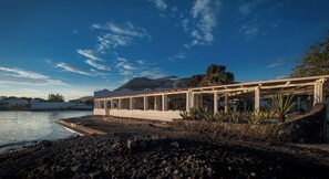A coastal building with a stone foundation, a flat roof, and a series of columns supporting a balcony. - Mari Del Sud Resort, Vulcano (Lipari)