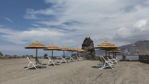 A beach with white lounge chairs and thatched umbrellas, a rocky formation, and a mountainous backdrop.