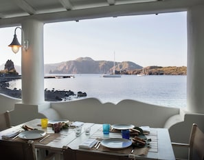 A dining table with plates, cutlery, and glasses set for two, overlooking a calm sea with boats and rocky terrain in the background. - Mari Del Sud Resort, Vulcano (Lipari)