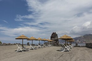 A beach with white lounge chairs and thatched umbrellas, a rocky formation, and a mountainous backdrop. - Mari Del Sud Resort, Vulcano (Lipari)
