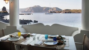 A dining table with plates, cutlery, and glasses set for two, overlooking a calm sea with boats and rocky terrain in the background.