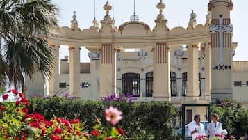 Un grand bâtiment somptueux, orné de nombreuses coupoles et de colonnes, entouré d’une végétation luxuriante et de fleurs aux couleurs vives.