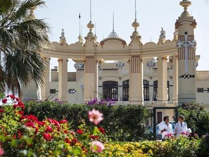 Un grand bâtiment somptueux, orné de nombreuses coupoles et de colonnes, entouré d’une végétation luxuriante et de fleurs aux couleurs vives.