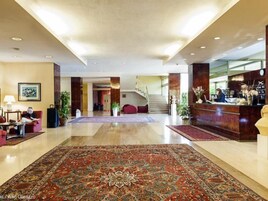 Um hall amplo com um tapete grande e ricamente decorado, um balcão de recepção e uma escadaria para os andares superiores.