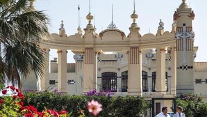 Un grand bâtiment somptueux, orné de nombreuses coupoles et de colonnes, entouré d’une végétation luxuriante et de fleurs aux couleurs vives.