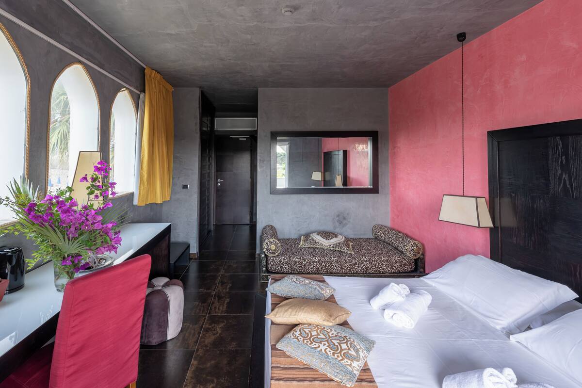 Una camera d'albergo moderna con un letto, un divano, una scrivania con un vaso di fiori e uno specchio grande.