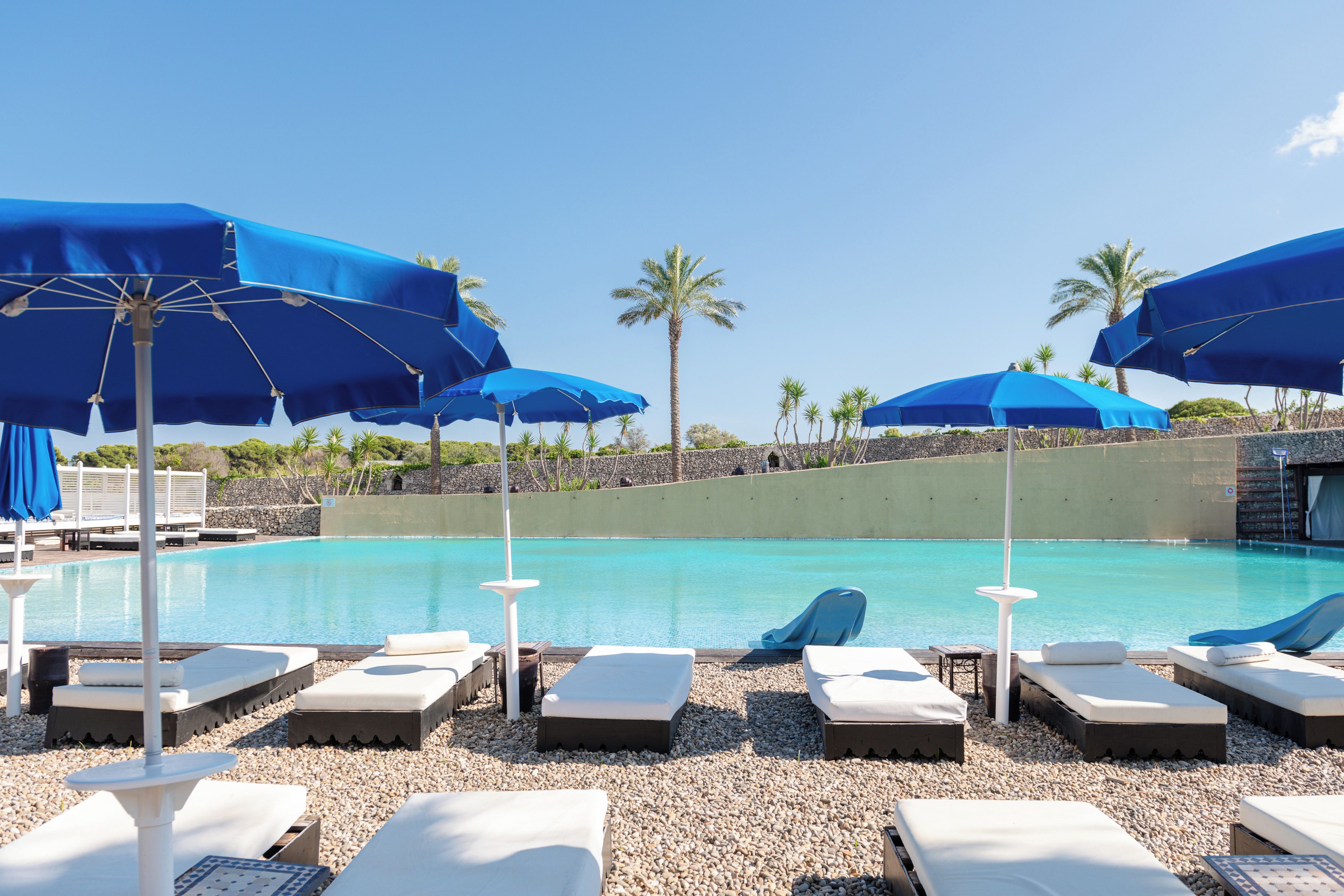 A poolside area with lounge chairs and blue umbrellas, surrounded by palm trees and a clear sky.
