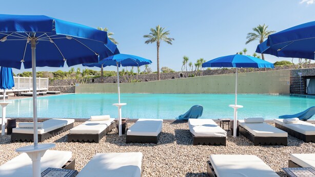 A poolside area with lounge chairs and blue umbrellas, surrounded by palm trees and a clear sky. - Amareclub Baia Dei Turchi Resort - Adults Only (Otranto)