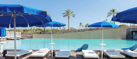 A poolside area with lounge chairs and blue umbrellas, surrounded by palm trees and a clear sky.