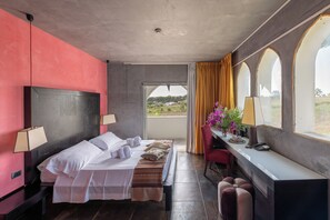 Quarto de hotel com cama, abajures, escrivaninha com cadeira, janela com cortinas e vista para o verde lá fora.