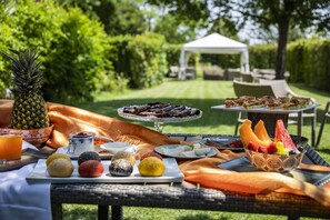 Een buiten eethoek met diverse soorten eten, zoals gebak, fruit en desserts, gepresenteerd op een tafel met uitzicht op een tuin en een gazebo op de achtergrond.