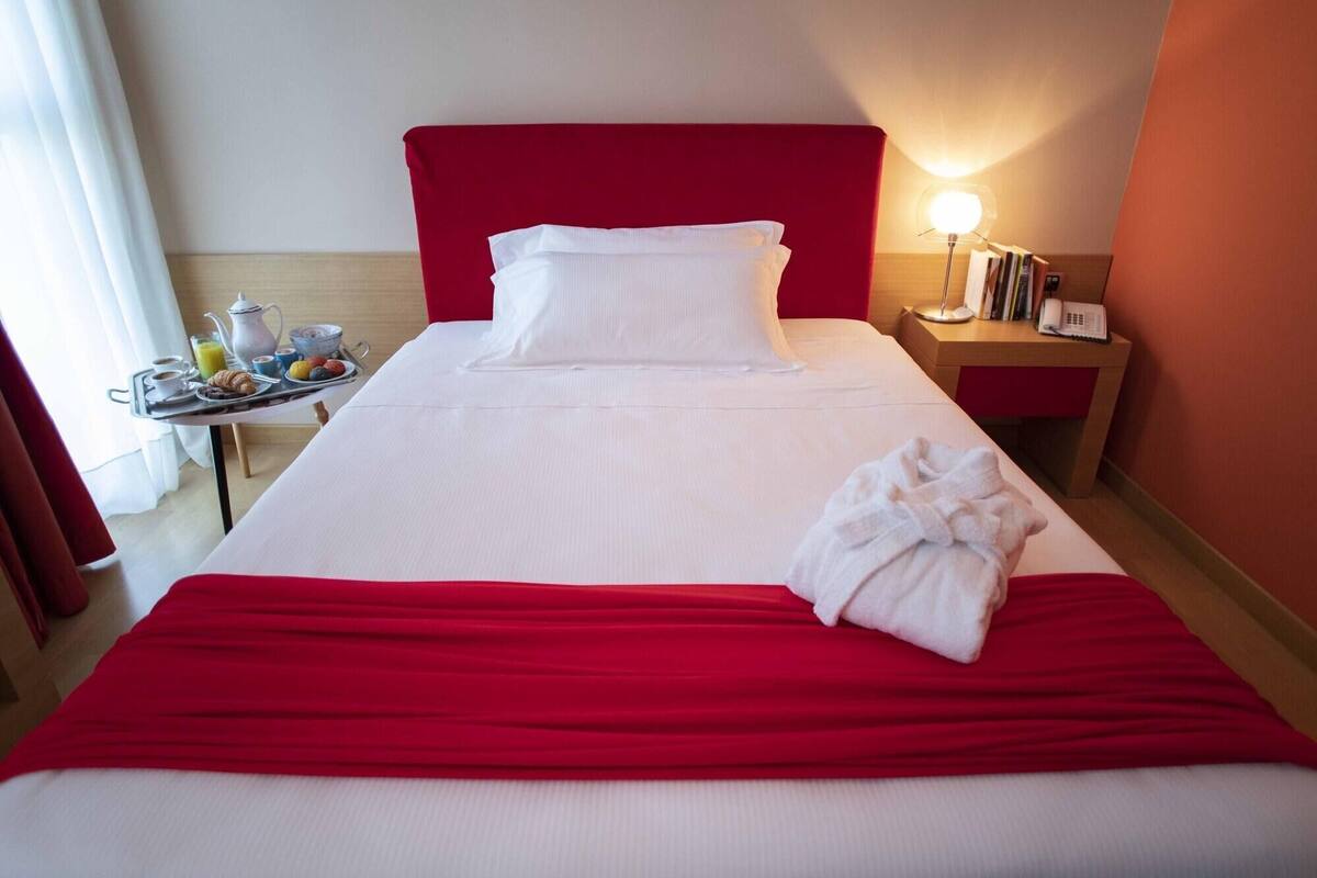 Une chambre d’hôtel avec un lit, une table de chevet avec une lampe, un plateau avec des articles pour le petit-déjeuner et une tête de lit rouge.