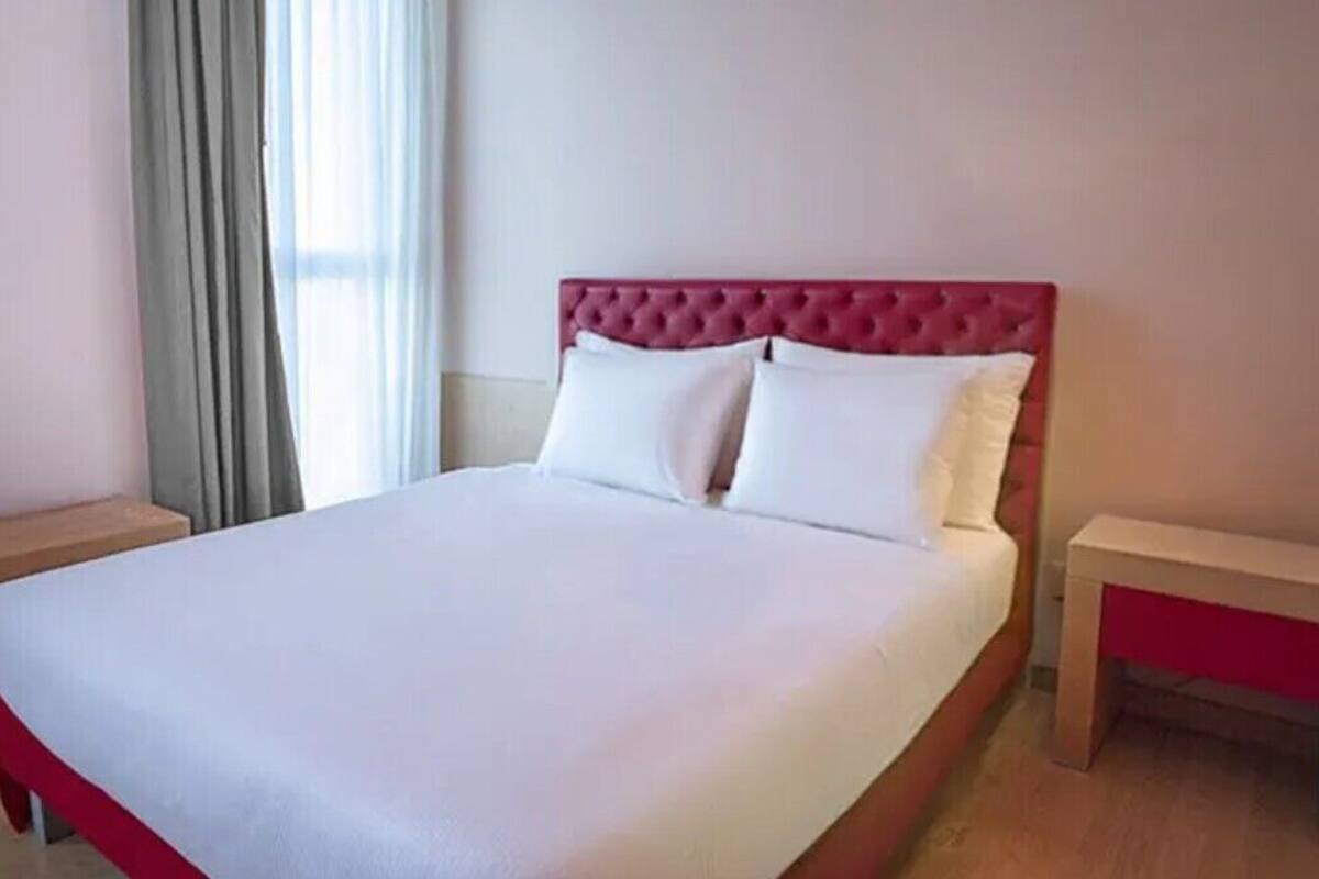 Un lit bien fait, avec une tête de lit rouge, deux tables de chevet et une fenêtre avec des rideaux.