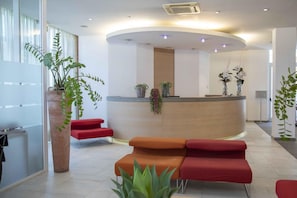 A modern reception area with a curved desk, red and orange seating, and potted plants. - B&B Hotel Remilia (Bibbiano)