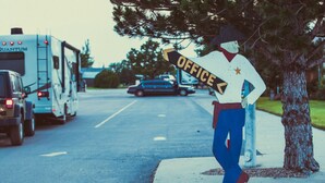 A person in a costume holding a sign that says "OFFICE" on a street with parked vehicles. - Amarillo by Morning (Amarillo)