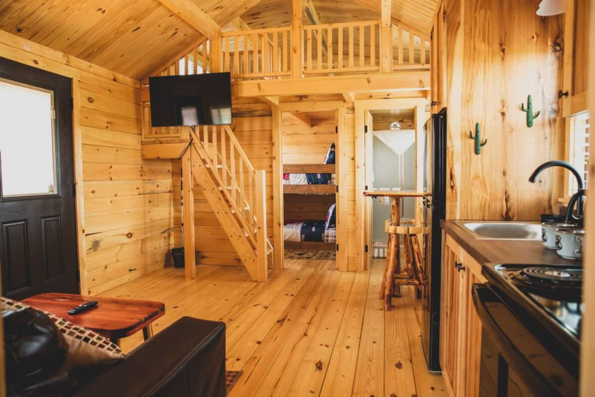 Interior de una cabaña de madera con cocina, comedor y una escalera que sube a un dormitorio en un altillo.