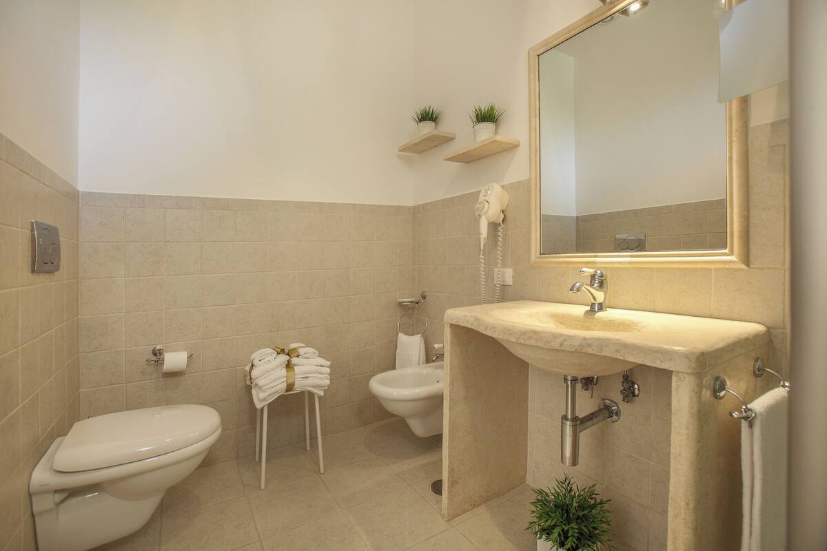 Ein Badezimmer mit Toilette, Bidet, Waschbecken und Spiegel. Darüber befinden sich zwei kleine Pflanzen auf Regalen.
