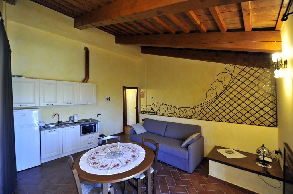 Une cuisine de taille réduite avec des meubles blancs, une table à manger recouverte d’une nappe à motifs floraux et un canapé gris.