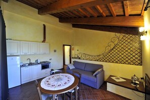 A compact kitchen with white cabinets, a dining table with a floral tablecloth, and a gray sofa. - Il Podere del Germano Reale (Coriano)