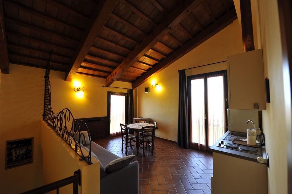 A cozy room with a wooden ceiling, a dining area with a table and chairs, a small kitchenette, and a staircase with a wrought iron railing.