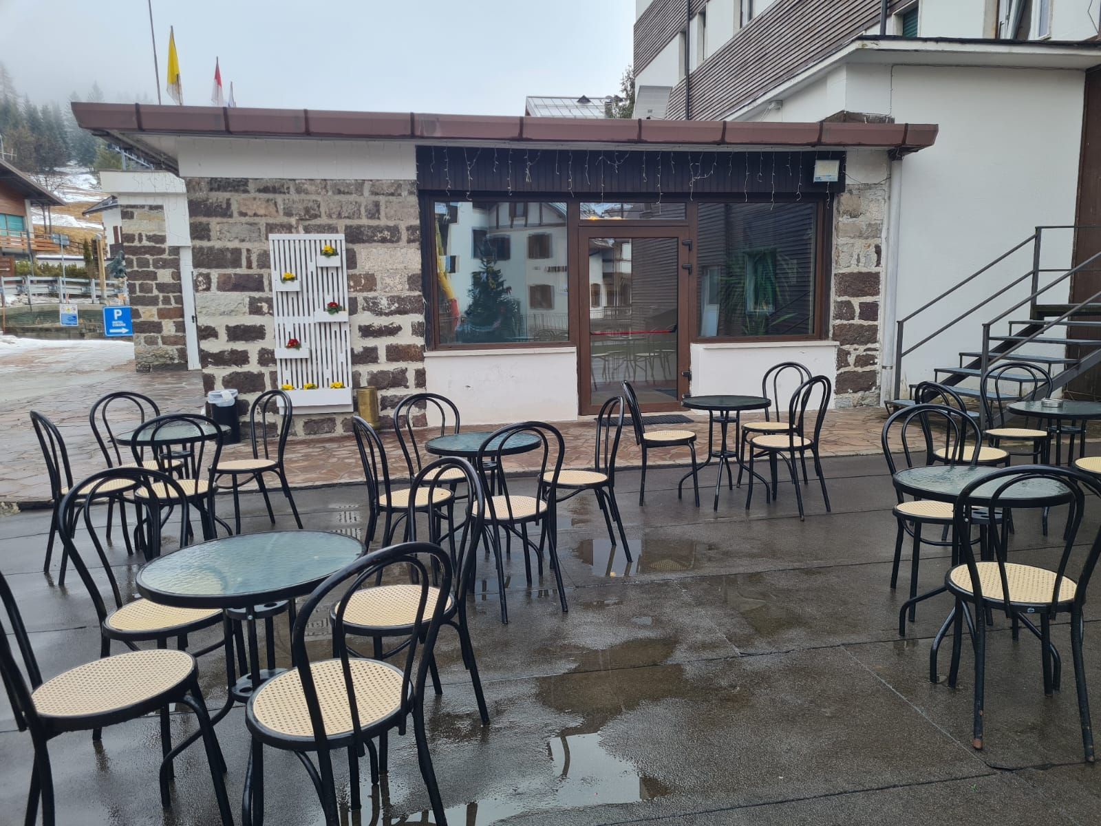 Un café al aire libre con mesas y sillas, un muro de piedra y una entrada con puerta de vidrio.