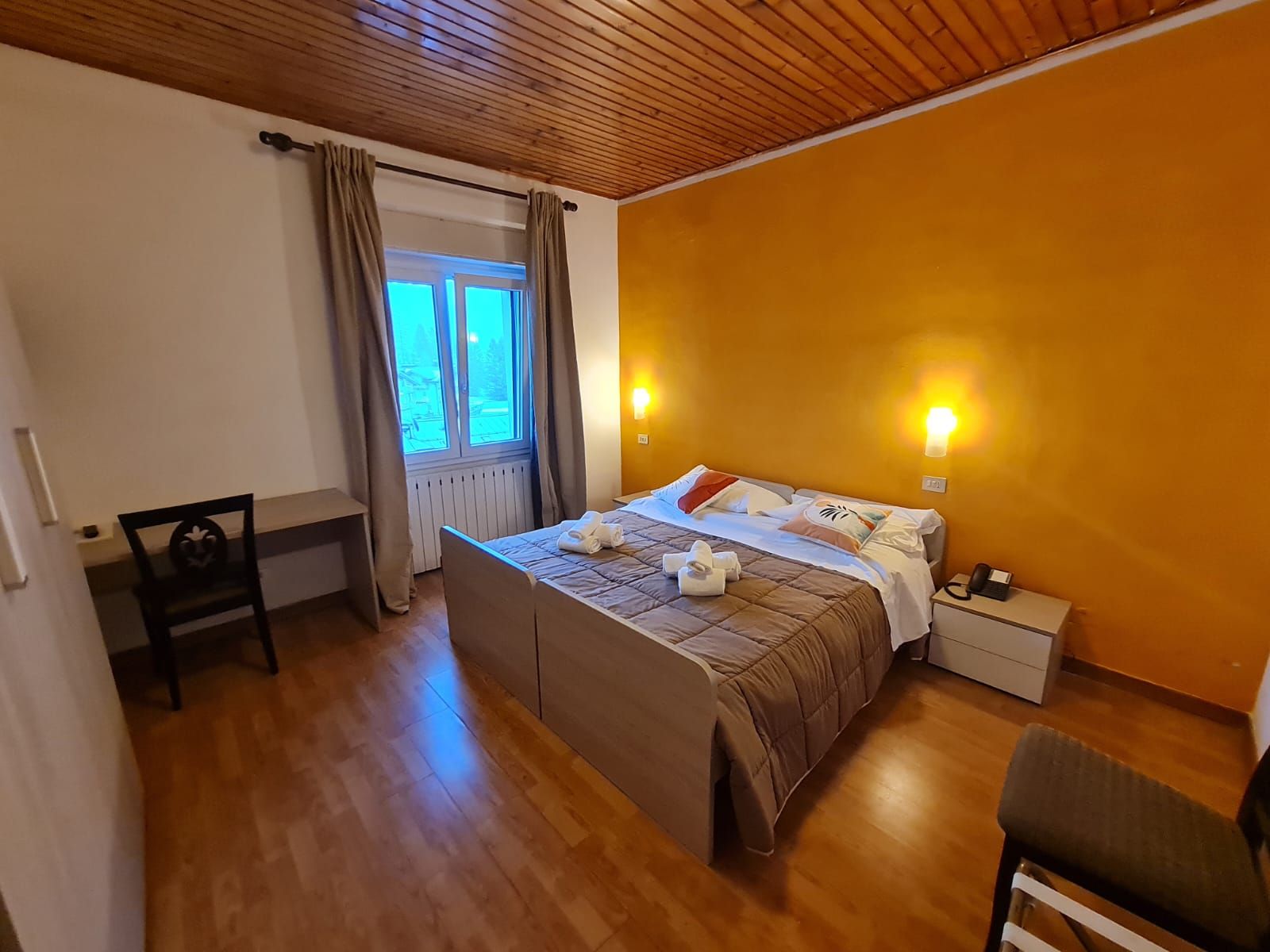 Una camera da letto con un letto, comodini, una sedia, una finestra con tende e un soffitto in legno.