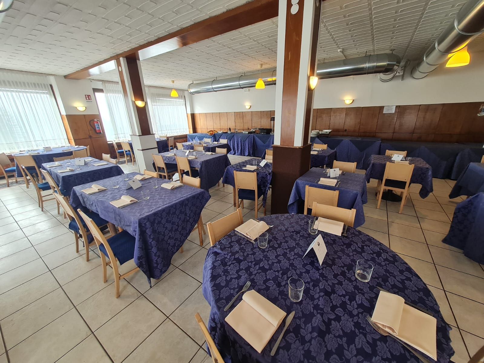 Zona pranzo con tavoli apparecchiati per gli ospiti, sedie in legno e pavimento piastrellato.