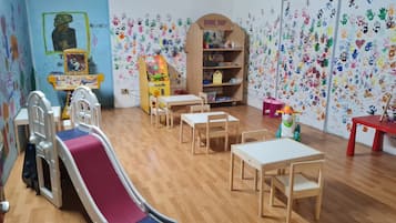 Un'area giochi per bambini con uno scivolo, tavoli, sedie, una macchinina e una libreria.