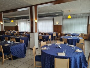 Een eetgedeelte met ronde tafels, bedekt met blauwe tafelkleden, houten stoelen en gele hanglampen.