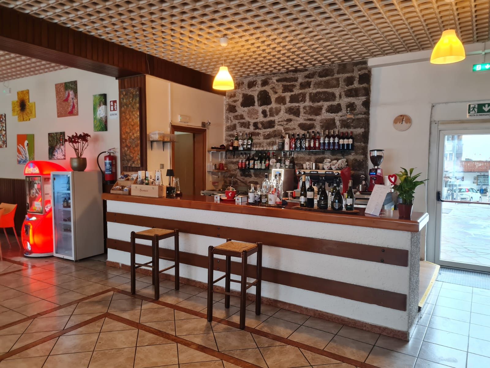 Zona bar con sgabelli, un bancone con diverse bottiglie, un estintore e opere d'arte alle pareti.