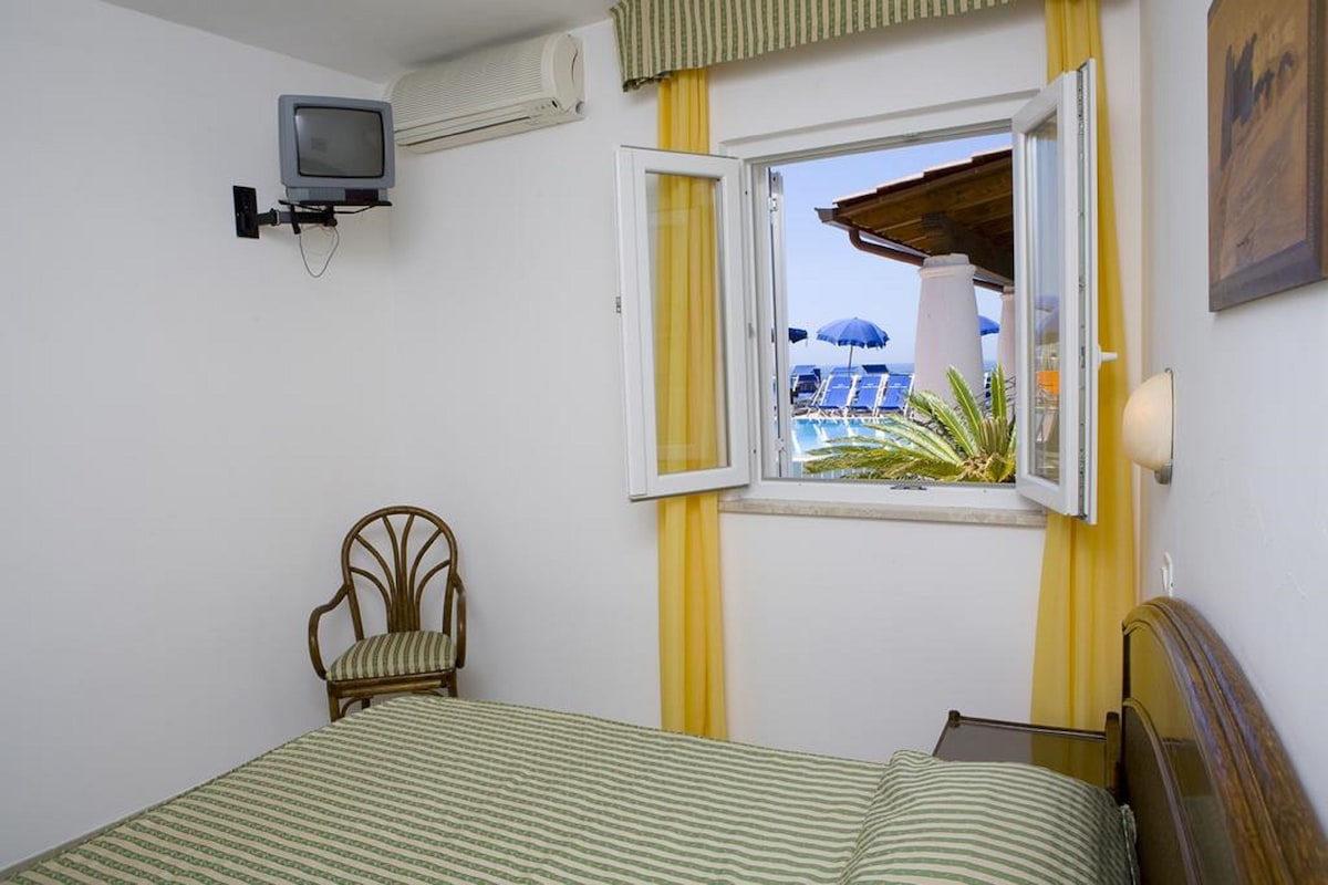 A room with a bed, a chair, a television, and a window with an open view of a pool and umbrellas.