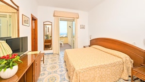 A hotel room with a bed, a desk with a TV, a mirror, and a view of the sea through a door. - Tramonto D'oro Terme (Forio)