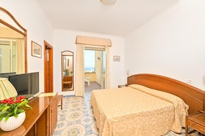 A hotel room with a bed, a desk with a TV, a mirror, and a view of the sea through a door. - Tramonto D'oro Terme (Forio)