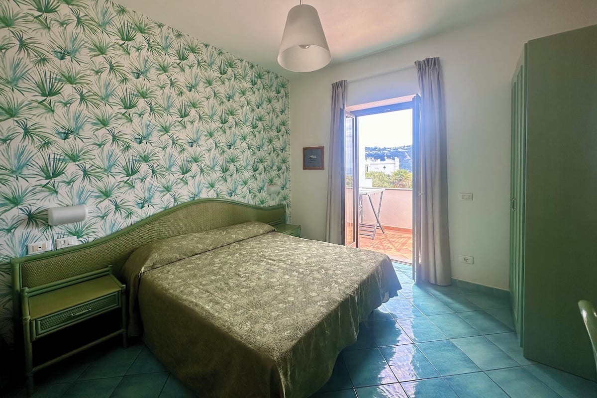 A room with a bed, a small table, a chair, a window with curtains, and a view of the outside.