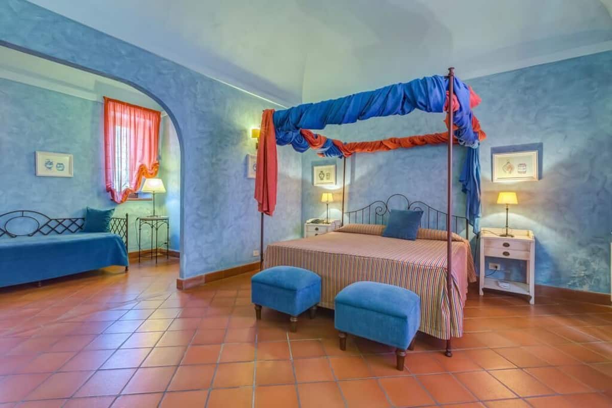 Quarto com cama de dossel, dois pufes azuis, mesa de cabeceira com abajur e sofá azul.