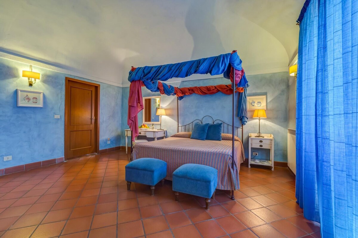 Un dormitorio con una cama con dosel, dos mesitas de noche, un velador, una ventana con cortinas y una puerta.