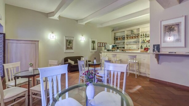 A dining area with a glass table, white chairs, and a vase of flowers. There is a kitchen area with shelves of bottles and a bar counter in the background. - Casa Delle Monache Boutique Resort (Castiglione di Sicilia)