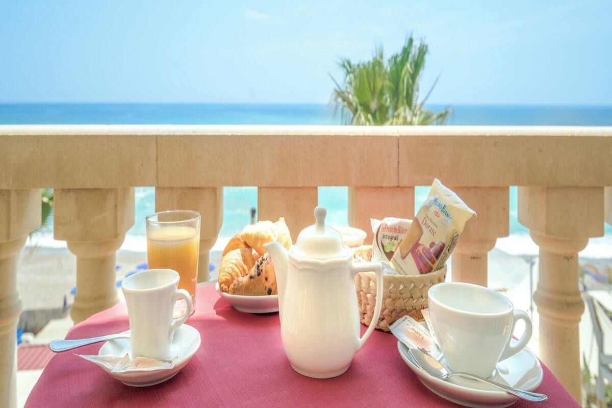 Ein Tisch mit Frühstücksangeboten, einer Karaffe und einer Tasse, vor dem Hintergrund eines Balkons mit Meerblick.
