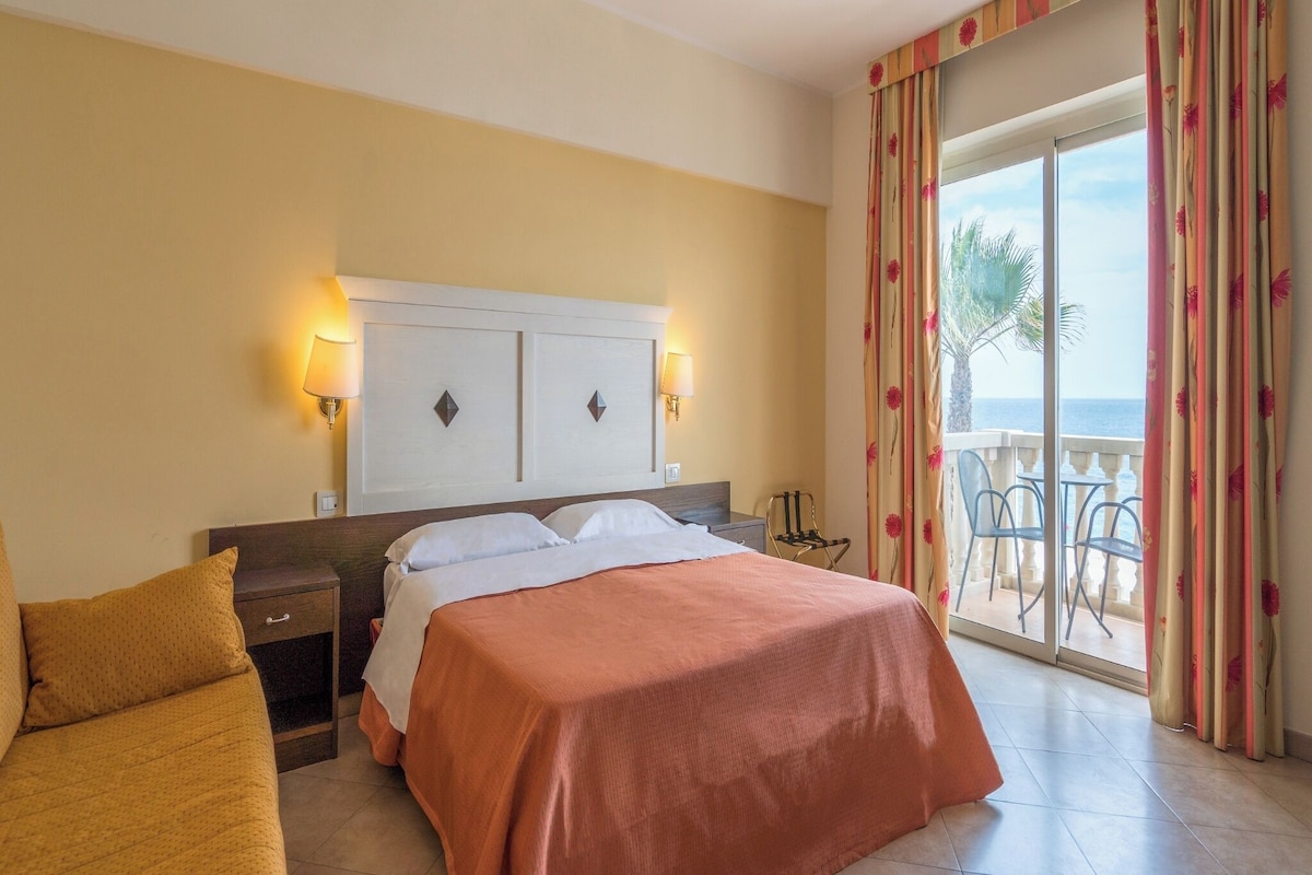 A hotel room with a bed, a sofa, a nightstand, a lamp, a window with curtains, and a view of the outdoors.