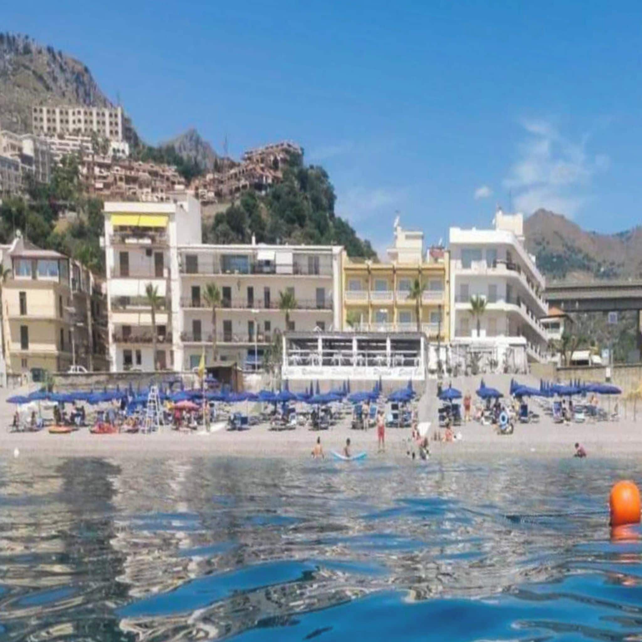 Un hotel sulla costa con diversi piani, una spiaggia attrezzata con ombrelloni e una vista libera sul mare.