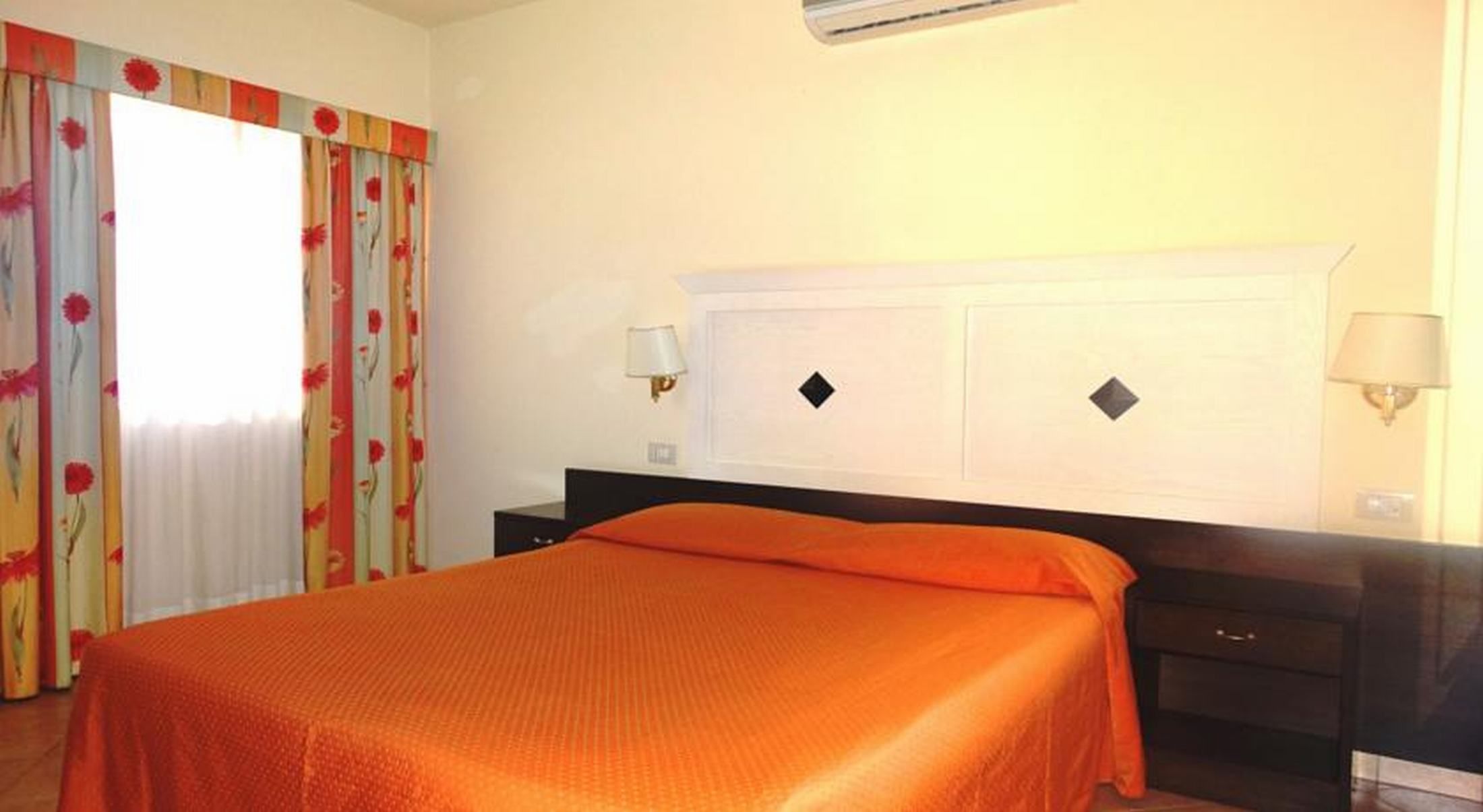 Una camera d'albergo con un letto, un comodino, una lampada e una finestra con tende.