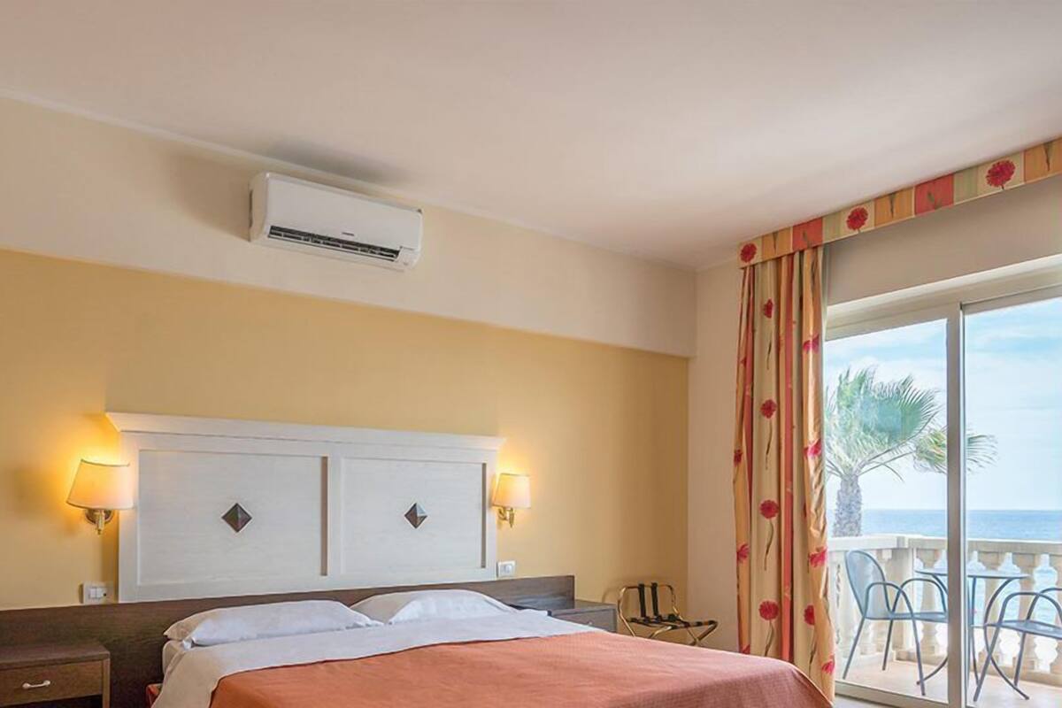 Ein Hotelzimmer mit Bett, zwei Nachttischlampen, einer Klimaanlage, einem Balkon mit Stühlen und Meerblick.