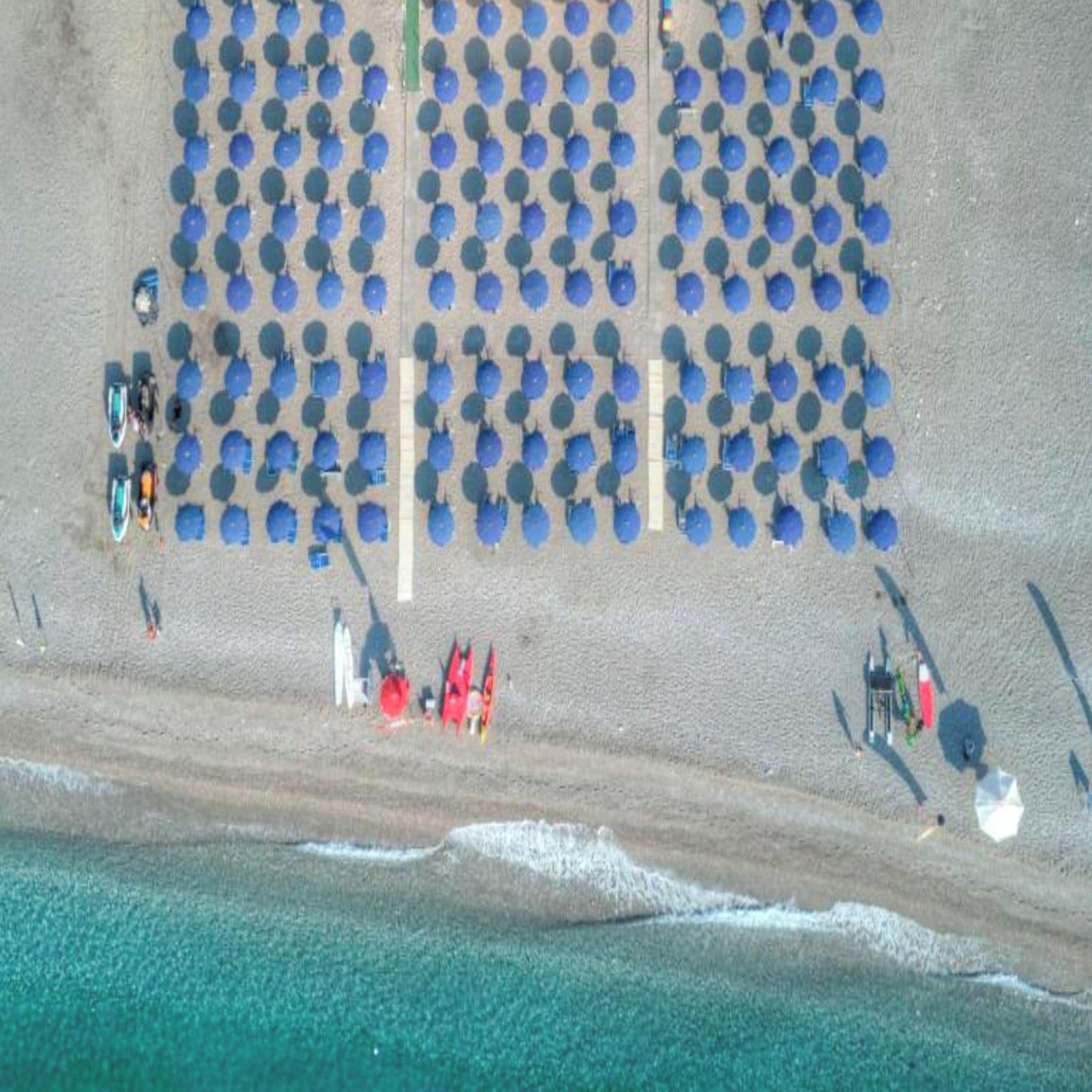 Une vue aérienne d’une plage avec des rangées de parasols bleus et de chaises de plage, une plage de sable et l’océan.