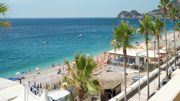A beach with clear blue water, palm trees, and a distant mountain. - Hotel Orizzonte (Letojanni)