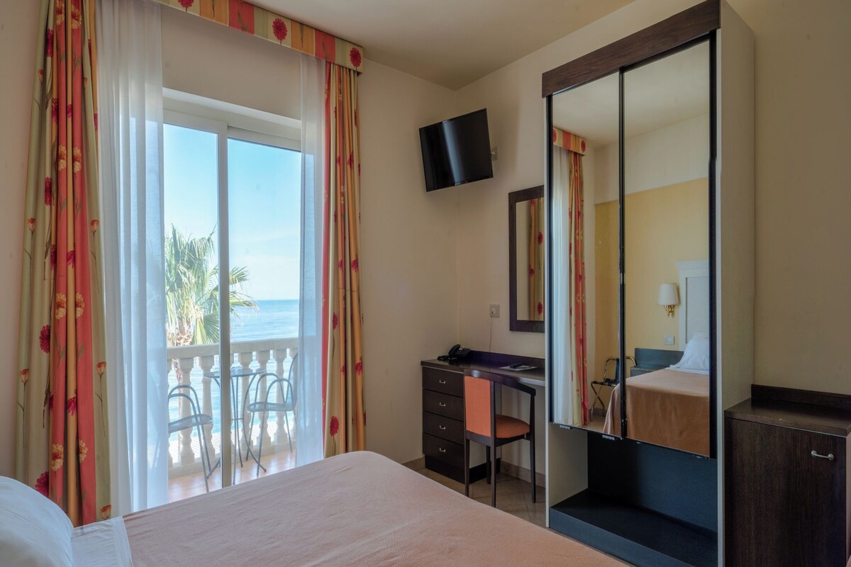 A hotel room with a bed, a desk, a wardrobe, a television, and a balcony with a view of the sea.