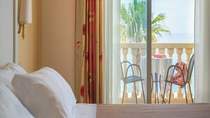 A hotel room with a bed, a nightstand, a lamp, a balcony with a table and chairs, and a view of palm trees and the sea. - Hotel Orizzonte (Letojanni)