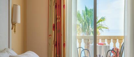 Un cuarto de hotel con una cama, una mesita de noche, una lámpara, un balcón con mesa y sillas, y vistas a palmeras y al mar.