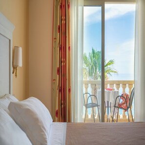 A hotel room with a bed, a nightstand, a lamp, a balcony with a table and chairs, and a view of palm trees and the sea. - Hotel Orizzonte (Letojanni)